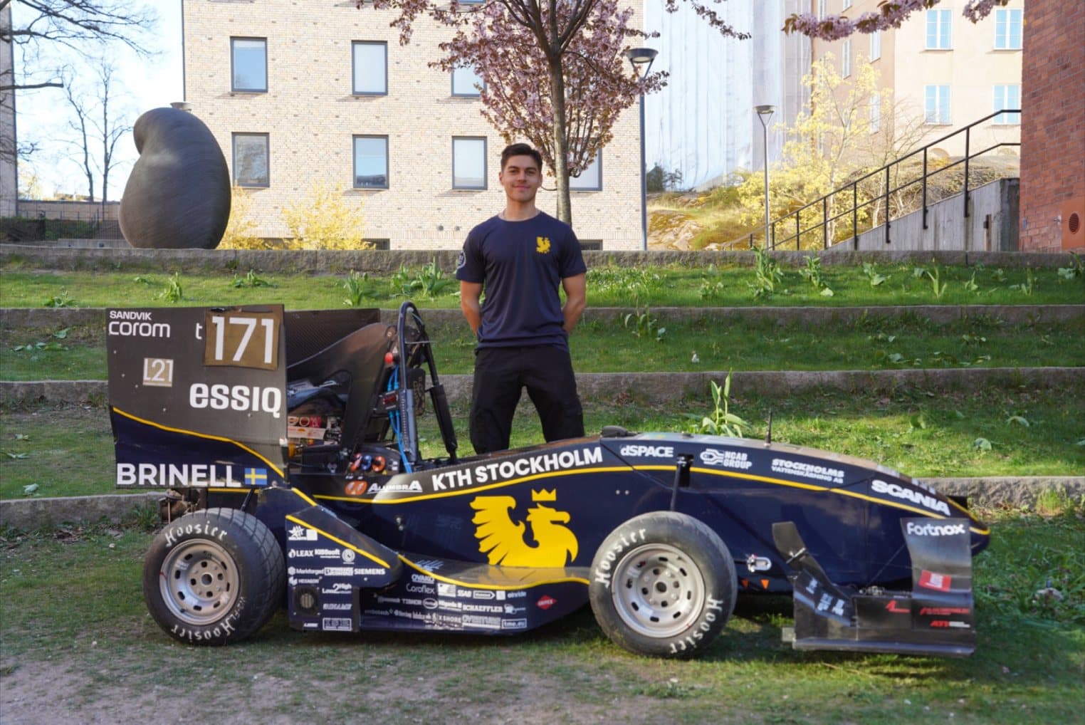 Adrian står bakom KTH Formula Students svarta och guldfärgade formelbil på gräset utanför KTH.
