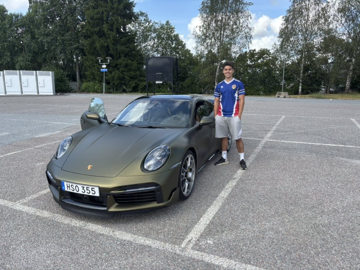 Adrian i fotbollströja bredvid en grön Porsche 911 på en parkering.