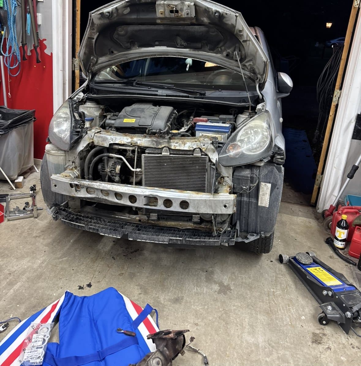 Grå småbil i ett garage med stötfångaren borttagen och motorrummet öppet.