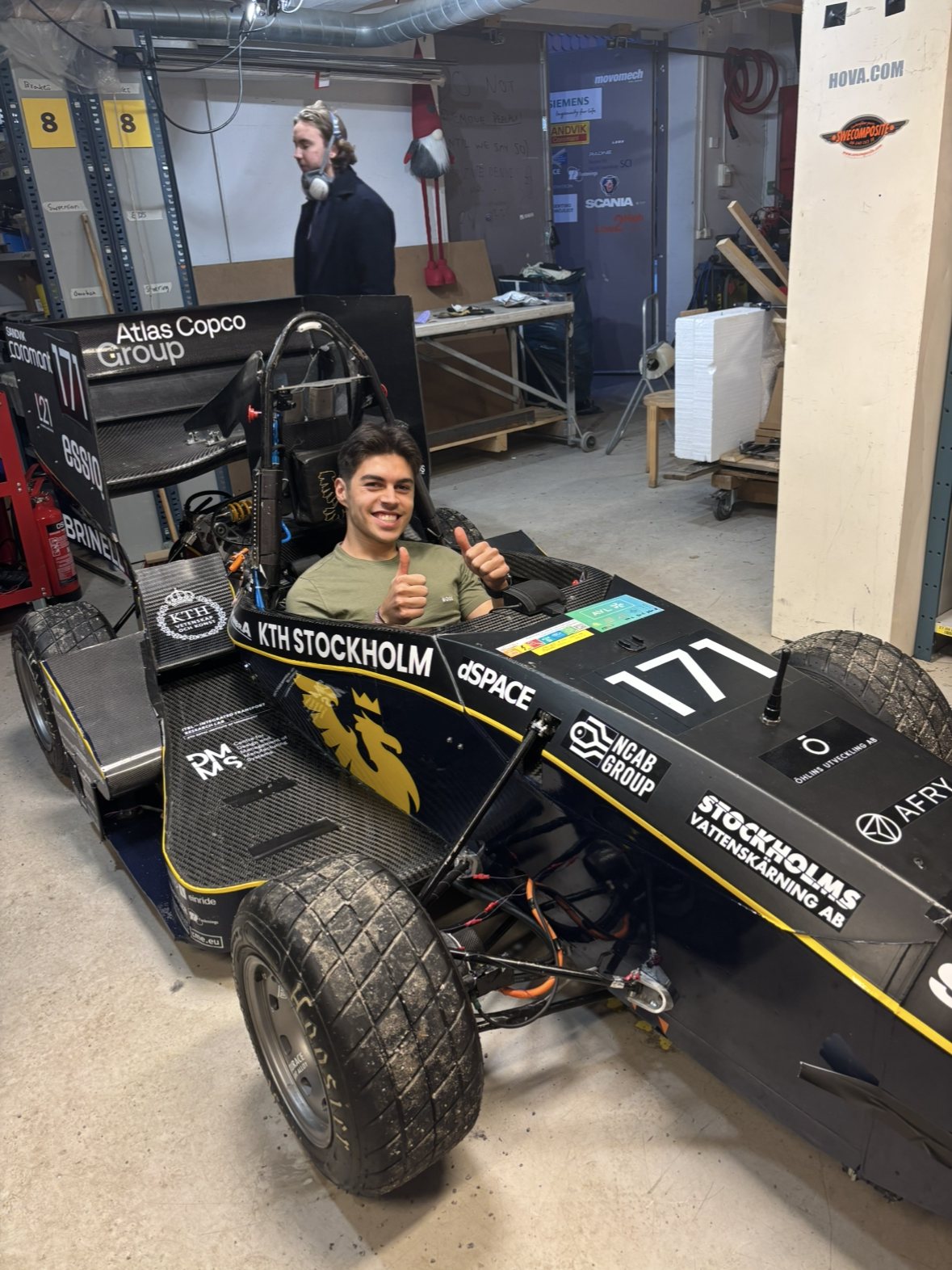 Adrian sitter i KTH Formula Students bil i verkstaden och gör tummen upp.
